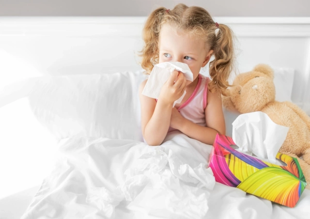 Kislány orrot fúj az ágyban, mellette plüss maci